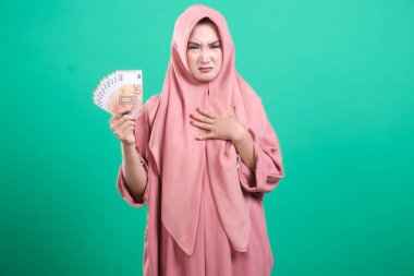 Tesettürlü hoşnutsuz bir Müslüman kadın, avro banknotlarını elinde tutarken, yeşil bir arka planda izole edilmiş, stüdyo çekimlerindeki mali stresi gösterirken hayal kırıklığı yaşıyor.