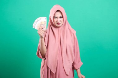 Başörtüsü takan bir kadın, yeşil arka planda izole edilmiş mali gerginliği gösteren hoşnutsuz bir ifadeyle avro banknotları tutuyor. Bu ekonomik zorlukları ve mali kaygıları temsil ediyor.