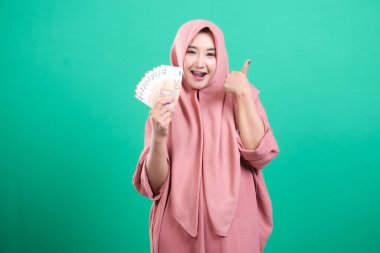 Yeşil arka planda izole edilmiş mali zaferden duyduğu mutluluğu aktaran, avro banknotlarını tutarken başparmak kaldıran tesettürlü Müslüman bir kadın. Geleceği için çok heyecanlı..