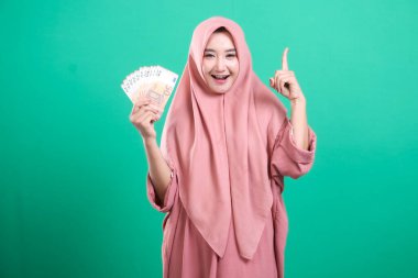 Elinde nakit para ve Eureka pozu olan tesettürlü genç Müslüman kadın, yeşil bir arka planda izole edilmiş mali başarısını ve zenginliğini gösteriyor. Yeşil arka planda servetiyle başarılı iş yatırımları sergiliyor..