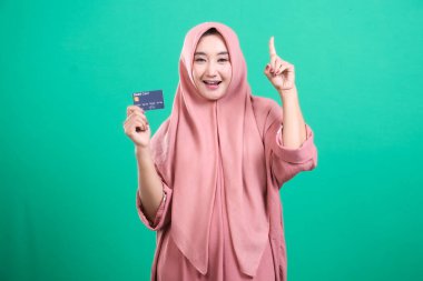 Kendine güvenen, tesettürlü, kredi kartı gösteren ve parlak bir fikir olduğunu gösteren genç bir kadın. Yeşil bir zemin üzerinde izole edilmiş, finansal yaratıcılık ve yenilik göstergesi.