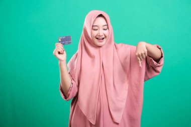 Tesettürlü neşeli müslüman kadın bir kredi kartı sunuyor ve parmağıyla aşağıya doğru işaret ediyor. Yeşil arka planda izole edilmiş, canlı bir zemin üzerinde modern kolaylık ve finansal fırsat sinyalleri veriyor..