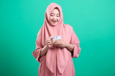 Türbanlı neşeli Müslüman bir kadın, yeşil bir arkaplanda izole edilmiş mali refah ve başarıyı sembolize eden Avrupa banknotlarını elinde tutuyor. Yeşil arka planda izole edilmiş parasal fırsatlar ve ekonomik büyümeyi somutlaştırıyor..