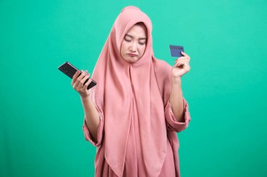 Elinde kredi kartı ve cep telefonu olan tesettürlü üzgün bir kadın, yeşil bir arka planda izole edilmiş mali sıkıntılar veya online alışveriş sorunları gösteriyor..