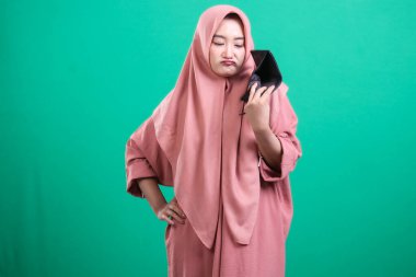 Başörtüsü takan genç bir kadın, yeşil bir arka planda izole edilmiş, maddi sorunları ve parasızlığı temsil eden boş cüzdanından duyduğu üzüntüyü ve hayal kırıklığını dile getiriyor..