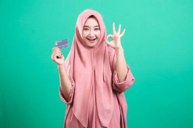Modern Müslüman bir kadının canlı portresi tesettürlü, onaylayıcı bir jestle bir banka kartı sunuyor, yeşil arka planda izole edilmiş bir mali güç olduğunu gösteriyor. Bankacılık ve çevrimiçi dükkanlarda çeşitli temsilleri resmetmek için ideal