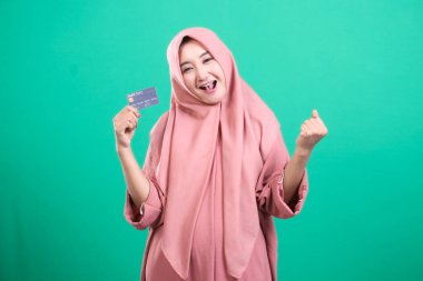 Tesettürlü neşeli Müslüman bir kadın yeşil bir arka planda izole edilmiş bir şekilde kredi kartını heyecanla gösteriyor. Güven saçıyor ve modern bankacılığı kucaklıyor..