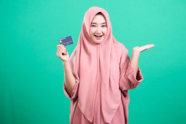 tesettürlü gülümseyen müslüman bir kadın kredi kartı tutuyor, olumlu bir jest sunuyor, yeşil bir arkaplanda izole edilmiş, uygun işlemleri sembolize ediyor ve finansal çözümleri güvence altına alıyor..