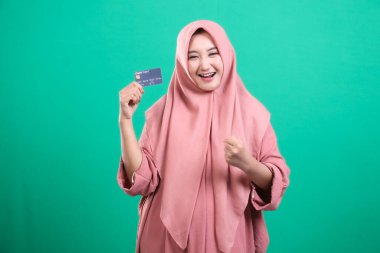 Bankamatik kartı olan mutlu genç Müslüman kadın yeşil arka planda izole edilmiş mali başarı ve iyi satın alma gücü hakkında heyecan ve sevinç ifade ediyor..