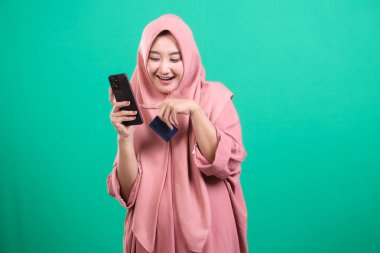 Tesettürlü neşeli Müslüman kadın telefon ve kredi kartı tutuyor, gülümserken ekranı işaret ediyor, yeşil arka planda izole edilmiş online alışverişin basitliğini gösteriyor..