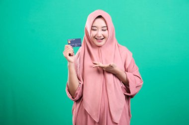 Elinde kredi kartıyla tesettürlü gülümseyen Müslüman kadın, dijital para ve rahatlık çağını gösteriyor, yeşil arka planda izole edilmiş, en az 90 karakter, en fazla 110 karakter.