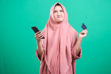 Genç Müslüman bir kadın, elinde telefon ve kredi kartı tutarken şaşkın görünüyor, muhtemelen mali zorluklarla karşı karşıya, yeşil bir arka planda izole edilmiş..