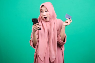Yeşil arka planda izole edilmiş, cep telefonu kullanan ve online alışveriş için kredi kartı tutan tesettürlü genç Müslüman bir kadın. Teknolojiyi ticaret için kullanan modern yaşam tarzı.