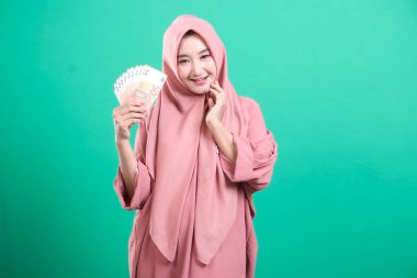 Avro banknotları tutan tesettürlü genç Müslüman kadın, yeşil bir arka planda izole edilmiş mali refah ve refahı sembolize ediyor. Başarı, zenginlik ve ekonomik bağımsızlığı temsil ediyor..