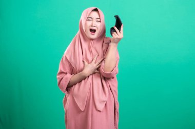 Başörtüsü takan genç bir müslüman kadın, şok geçiriyor ve yeşil arka planda izole edilmiş kırık bir ekranı olan kırık bir akıllı telefonu tutuyor. Mobil acil durum oldu..