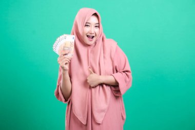 Avrupa banknotları tutan ve başarı ve zenginliği ifade eden, yeşil arka planda izole edilmiş, tesettürlü mutlu Müslüman kadın. Yeşil arka planda izole edilmiş mutluluk ve refahı gösteriyor..