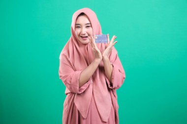 Başörtülü neşeli bir Müslüman kadın yeşil bir arka planda izole edilmiş, güvenli mali işlemleri savunan, kredi kartını gururla gösteriyor. Çağdaş bankacılık ve tüketiciliğin tanıtımı, stüdyo çekimi.