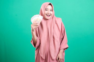 Mutlu bir ifadeyle avro banknotlarını tutan tesettürlü neşeli genç Müslüman kadın, yeşil arka planda izole edilmiş mali başarıyı sembolize ediyor.