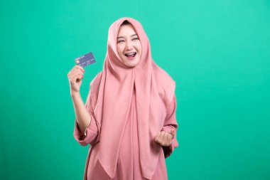 Bankamatik kartıyla pembe tesettürlü heyecanlı Müslüman kadın, yeşil bir arka planda neşeli bir şekilde izole edilmiş, finansal başarısını kutluyor..