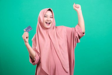 Neşeli Müslüman bir kadın, yeşil bir arka planda izole edilmiş koluyla bir kredi kartı tutarak başarısını ve finansal özgürlüğünü ifade ederek kutluyor..
