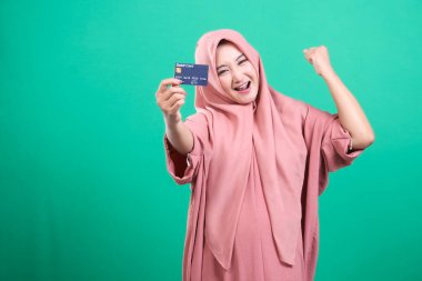 Zevkli bir Müslüman kadın banka kartını sergiliyor ve yeşil arka planda izole edilmiş mali refahı ve yetkiyi simgeleyen zaferini kutluyor..