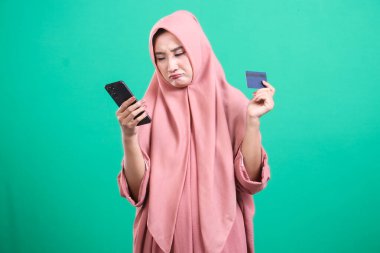 Akıllı telefonu ve kredi kartı somurtması olan hayal kırıklığına uğramış bir kadın, muhtemelen işlem hatası ya da mali sorunları yüzünden. Yeşil bir arka planda izole edilmiş. Parayla ilgili endişelerini ifade eden bir bayanın stüdyo fotoğrafı..