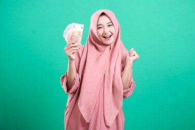 Başörtülü neşeli kadın, refah ve başarıyı sembolize eden, yeşil bir arka planda izole edilmiş, finansal başarısını temsil eden bir tomar avroyu neşeyle sergiliyor..