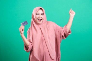 Tesettürlü neşeli Müslüman bir kadın, zafer dolu bir şekilde bir kredi kartı tutuyor ve elini kaldırıyor, finansal özgürlüğü temsil ediyor, yeşil bir arka planda izole edilmiş, başarıyı ve güçlendirmeyi simgeliyor, 97 karakter,.