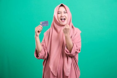 Başörtüsü içinde sevinçli bir kadın banka kartıyla kutlama yapıyor, yüzü yeşil arka planda izole edilmiş bir başarıdan sonra mutluluk saçıyor. Neşe ve modern finansal başarı sergiliyor..