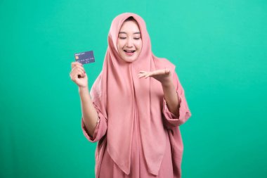 Türbanlı çekici Müslüman kadın, yeşil bir zeminde izole edilmiş, çevrimiçi ticaretin özünü yakalayan modern finansmanın gücünü gösteren bir banka kartı sunuyor. Reklam ve promosyon için ideal. Erişilebilirlik ve empoze sunuyor.