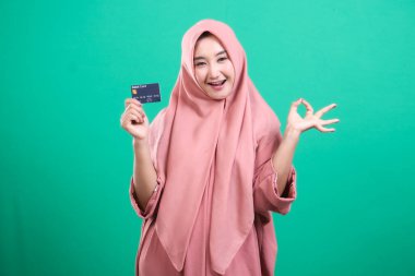 Başörtüsü takan mutlu Müslüman kadın bir kredi kartı tutuyor ve online bankacılıktan memnun olduğunu belirten bir işaret veriyor. Yeşil arka planda izole edilmiş, nakit ödeme ve finansal özgürlükleri gösteriyor..