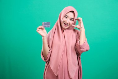 Yeşil bir arka planda izole edilmiş neşeli bir mutluluk ifadesiyle bankamatik kartını gösteren keyifli genç Müslüman kadın. Nakitsiz modern yaşam tarzının ve finansal yetkilendirmenin altını çiziyorum.