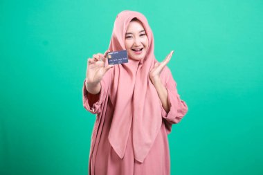 Başörtülü memnun Müslüman kadın, yeşil arka planda izole edilmiş, modern finans hizmetlerine ve ekonomik fırsatlara erişimi vurgulayan bir banka kartı sunuyor..