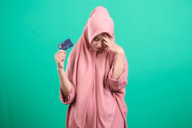 Kredi kartı tutan tesettürlü, tesettürlü Müslüman bir kadın ve mali durumla ilgili bir jest, yeşil bir arka planda izole edilmiş. Resim harcamalarda kasvetli bir yansımayı betimler.