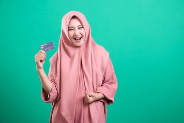 tesettürlü neşeli müslüman kadın yüzünde neşeli bir ifadeyle kredi kartı sergiliyor. Yeşil bir zemin üzerinde izole edilmiş, finans ve yaşam tarzı teması için ideal, izleyiciyi rahat ve fırsat dolu bir dünya hayal etmeye davet ediyor..