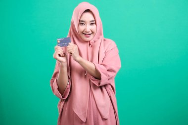 Genç tesettürlü, tesettürlü Müslüman bir kadın elinde bir kredi kartıyla, yeşil arka planda soyutlanmış modern İslami finans ve yaşam tarzını resmediyor. Finansal bağımsızlık ve dijital ödemeleri vurguluyor.