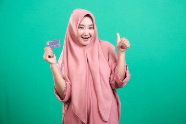 Neşeli Müslüman kadın, yeşil bir arka planda izole edilmiş, finans hizmetlerinden memnun olduğunu gösteren bir kredi kartı sunuyor..