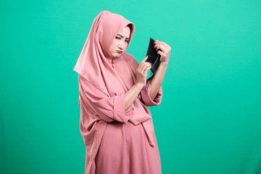 Başörtüsü takan genç bir kadın, ne yazık ki boş cüzdanına bakar. Yeşil bir arka planda izole edilmiş mali zorluklar ve endişeleri ifade eder..