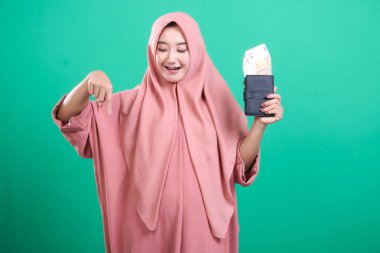 Pembe tesettürlü çekici Müslüman kadın yeşil arka planda izole edilmiş avro banknotlarıyla dolu bir cüzdanı sergiliyor. Görüntü finansal refahı ve kültürel takdiri simgeliyor..