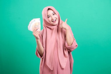 Tesettürlü mutlu bir Asyalı kadın yeşil arka planda izole edilmiş avro banknotlarının hayranı, somut mali başarısını sergiliyor. Bu resim mutluluğu ve gücü resmediyor, kazancı kutluyor..
