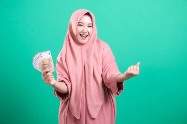 Elinde yeşil bir arka planda izole edilmiş avro banknotları tutan geleneksel tesettürlü heyecanlı genç Müslüman kadın. O, ekstra gelir aldıktan sonra neşeli ve mutlu..