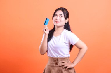 Genç bir kadın elinde bir kredi kartı tutarken düşünceli bir şekilde yukarı bakar, muhtemelen satın alma kararını düşünürken. turuncu arkaplanda izole