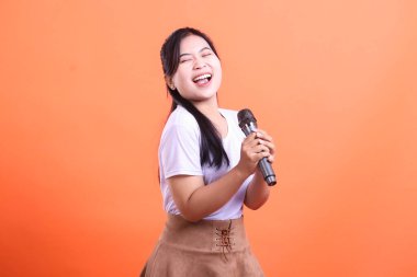 Bir kadın mikrofona mutlu bir şekilde şarkı söylüyor, gözleri kapalı ve geniş bir gülümseme turuncu arka planda izole edilmiş tutku ve neşe sergiliyor.