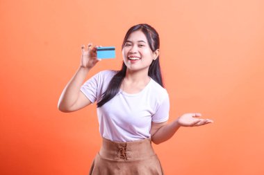 Genç bir kadın kredi kartını gösterirken gülümsüyor, görünüşe göre alışverişe hazır, kendine güveni ve neşesiyle, turuncu arka planda soyutlanmış.