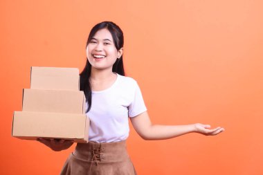Sırıtkan Asyalı kadın elinde bir yığın karton kutu tutuyor. Turuncu arka planda izole edilmiş müşteriye bir paket teslim etmeye ya da göndermeye hazır.