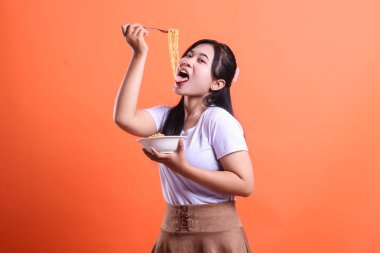 Çatalla bir kase erişte yiyen genç bir kadın, turuncu arka planda izole edilmiş lezzetli yemeği tatmak için dilini dışarı çıkarıyor..