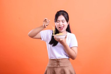 Neşeli genç bir kadın lezzetli bir erişte kasesinin tadını çıkarıyor, her lokmanın tadını çıkarıyor ve mutlu bir yüz ifadesiyle, turuncu arka planda izole edilmiş..