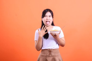 Eriştenin tadına baktıktan sonra iğrenç bir ifade sergileyen genç bir kadın elinde yemekle bir kase tutuyor. turuncu arkaplanda izole