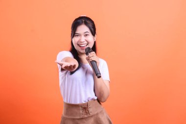 Mikrofonlu mutlu bir Asyalı şarkıcı sizi açık elleriyle davet ediyor ve turuncu arka planda izole bir şekilde gülümsüyor.