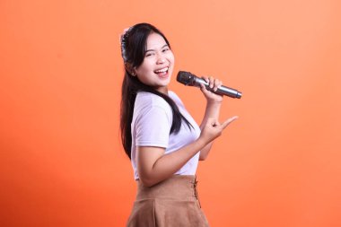 Gülümseyen genç bir kadın mikrofonu sanki şarkı söyler gibi tutuyor, diğer eliyle işaret ediyor. turuncu arkaplanda izole